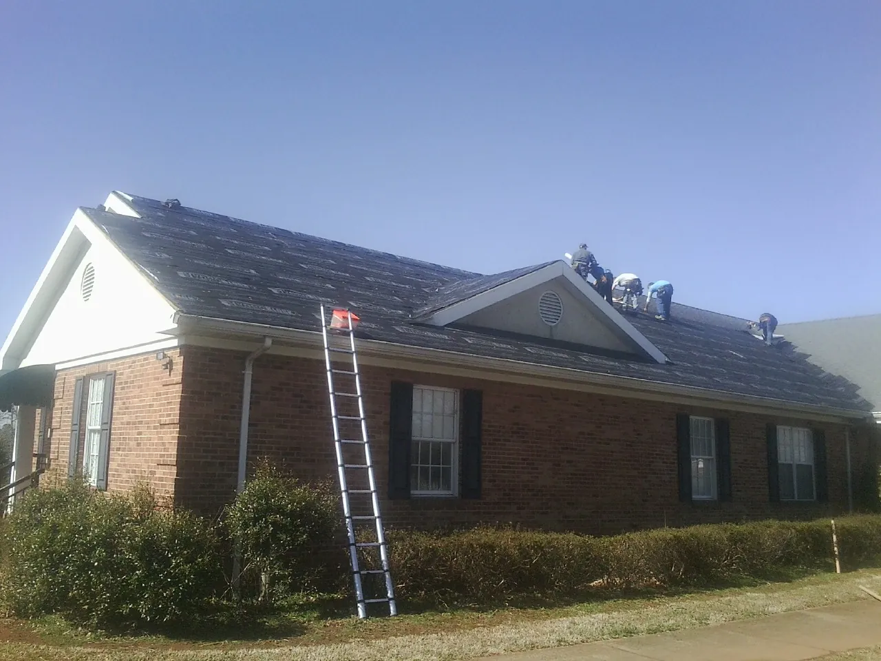Crew tearing shingles off a brick ranch roof with a ladder set — Emergency Roof Repair work by our crew in East Rockaway