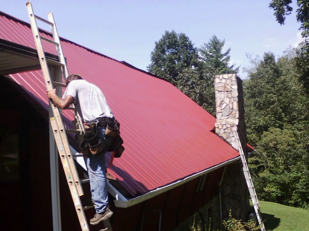 Roofer on a ladder beside a red metal roof and stone chimney — Soffit & Fascia Repair by Roofing Brothers in East Rockaway